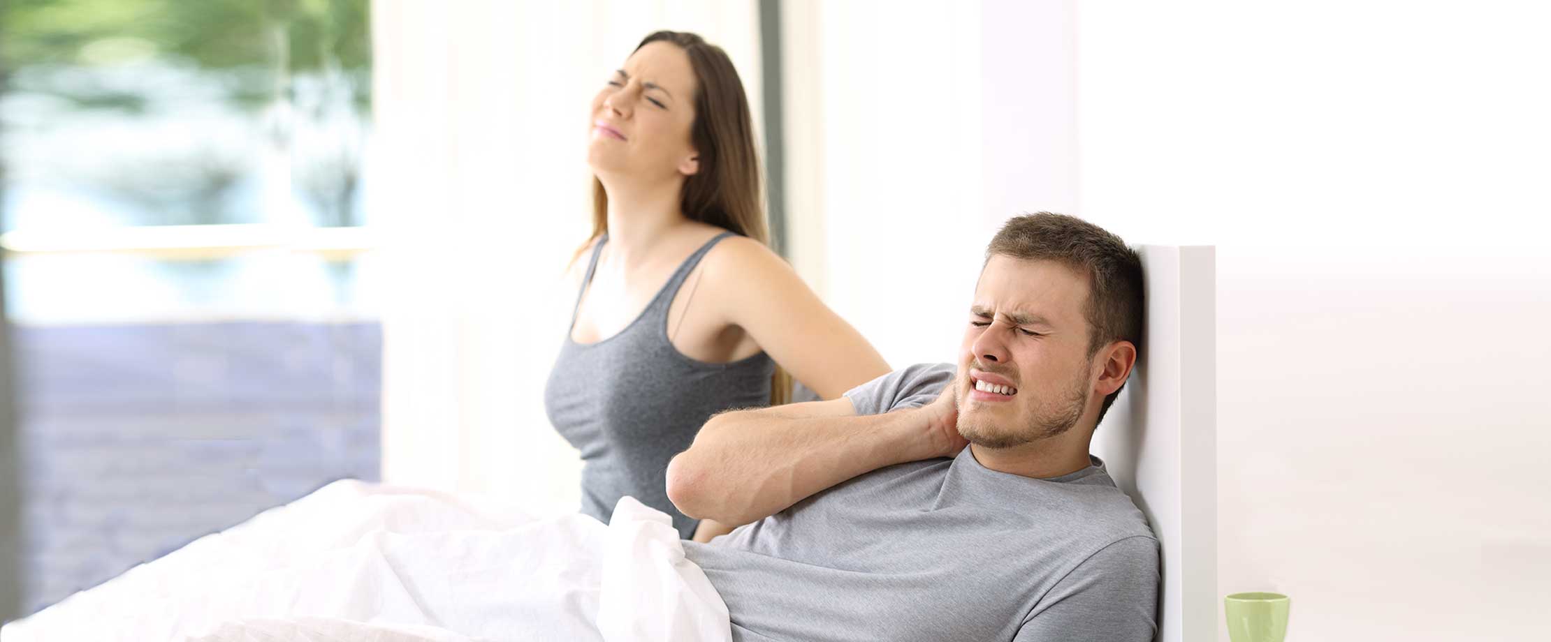 Ein Pärchen liegt morgens im Bett. Beide haben schmerzverzerrt das Gesicht verzogen und greifen sich auf den Rücken und das Genick, das zu schmerzen scheint.